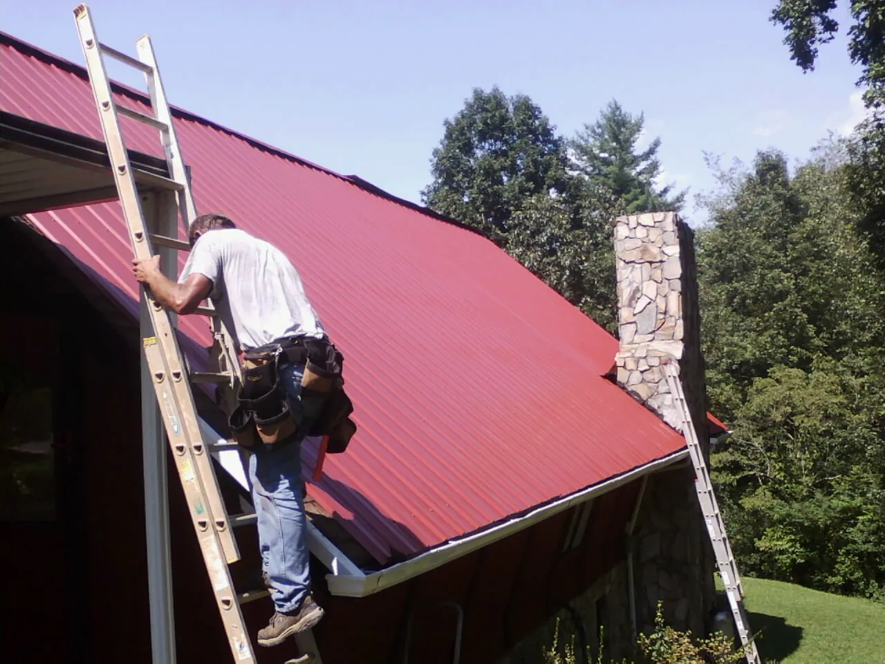 Roofer on a ladder beside a red metal roof and stone chimney — Soffit & Fascia Repair by Roofing Brothers in Madbury