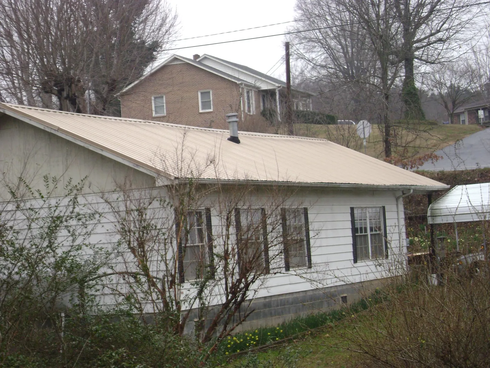 Tan metal roof on a white sided house set into a hillside — Commercial Roofing by Roofing Brothers in Madbury